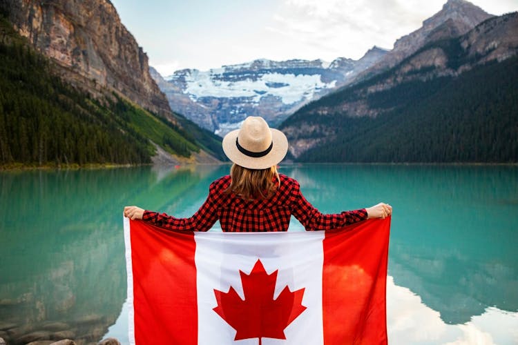 Immigration to Canada, Woman Wearing Red and Black Checkered Dress Shirt and Beige Fedora Hat Holding Canada Flag Looking at Lake